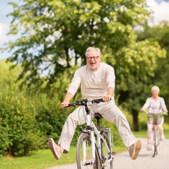 Idoso andando de bicicleta, ao fundo uma mulher também pedalando